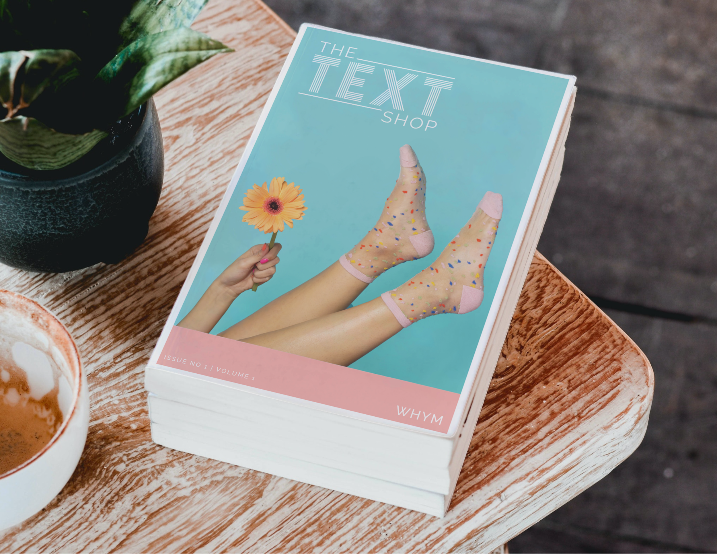 Mockup of The Text Shop catalog on a wooden table, cover showing legs in confetti socks and a hand holding a gerbera daisy